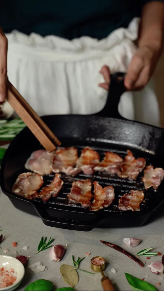 Sizzling bacon slices cooking in a cast iron pan with seasonings.