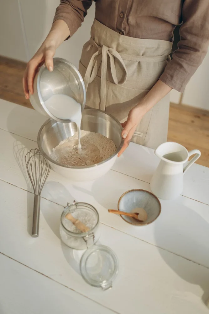 Close-up of a person pouring milk into flour in a mixing bowl, preparing dough.