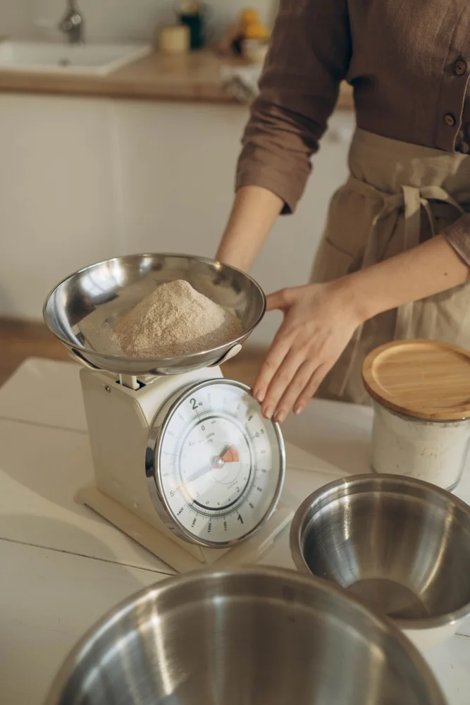 A woman weighing flour using a kitchen scale, preparing for baking in a cozy kitchen setting.
