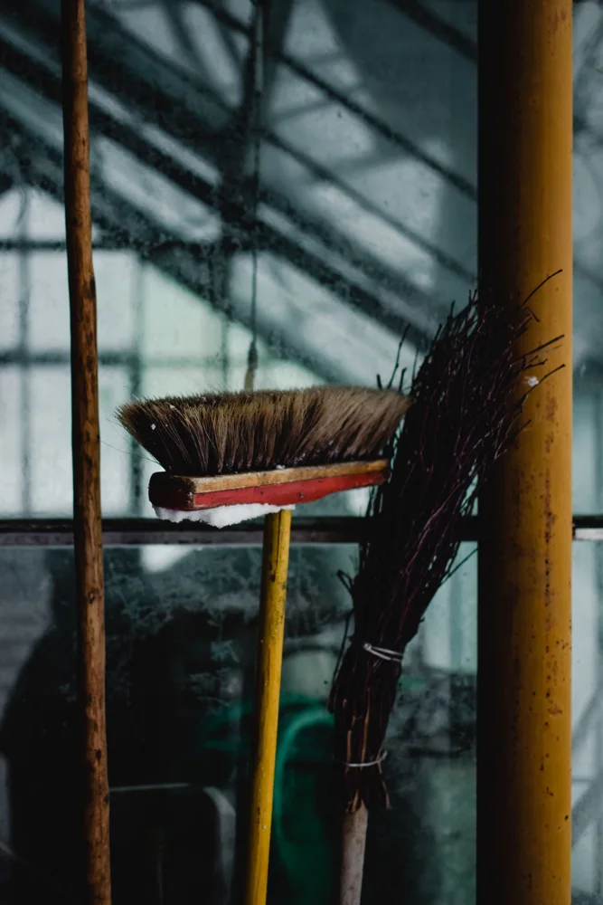 Set of brooms and cleaning tools leaning against a rustic glass wall.