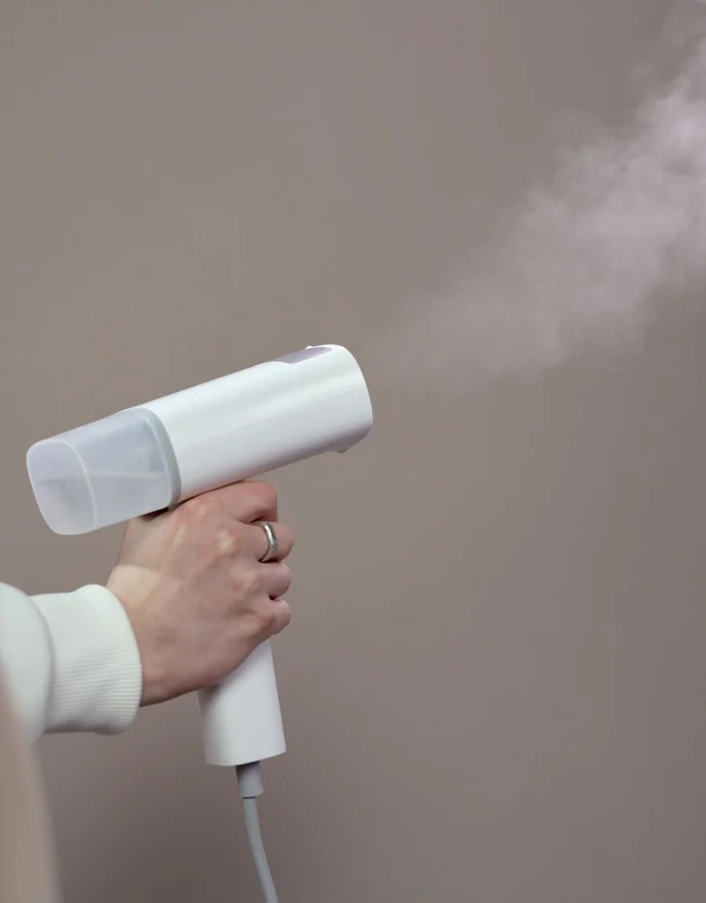 A close-up of a person using a handheld clothes steamer against a neutral background.