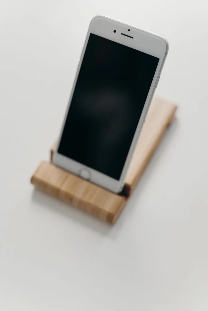 A minimalist image of a smartphone with a blank screen on a wooden stand against a plain background.