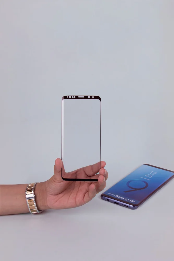 Close-up of a hand holding a tempered glass screen protector beside a smartphone.
