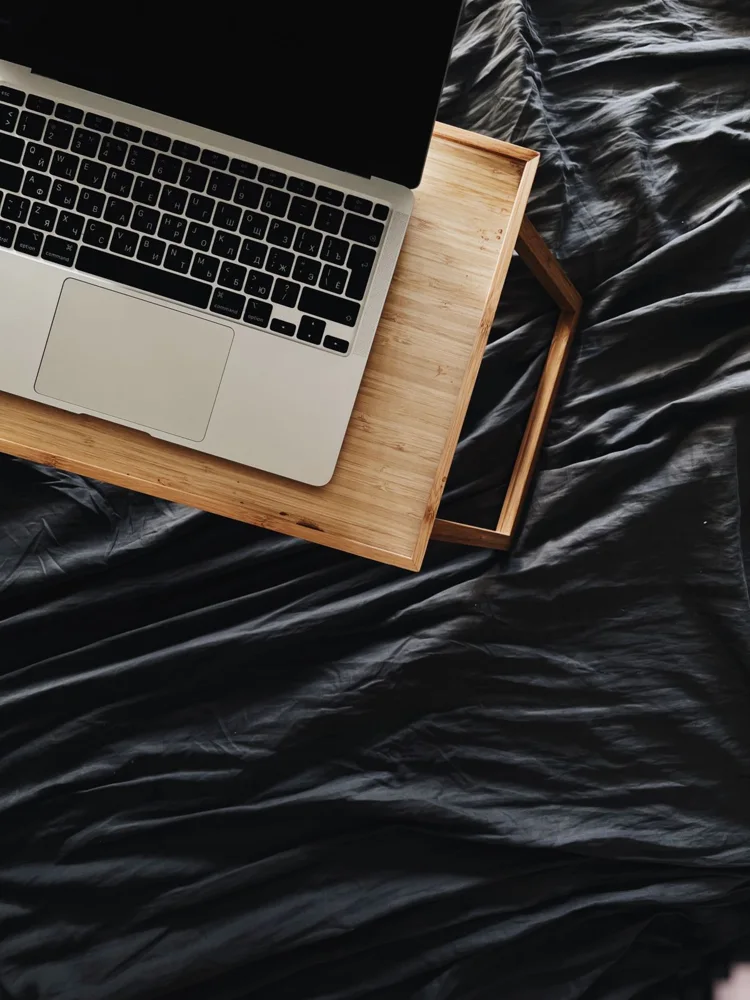 A laptop on a wooden stand resting on a dark bedspread, creating a cozy workspace.