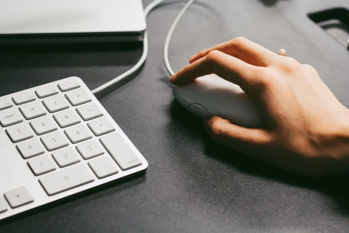 Hand operates a computer mouse beside a white keyboard on a dark desk, showcasing modern technology use.