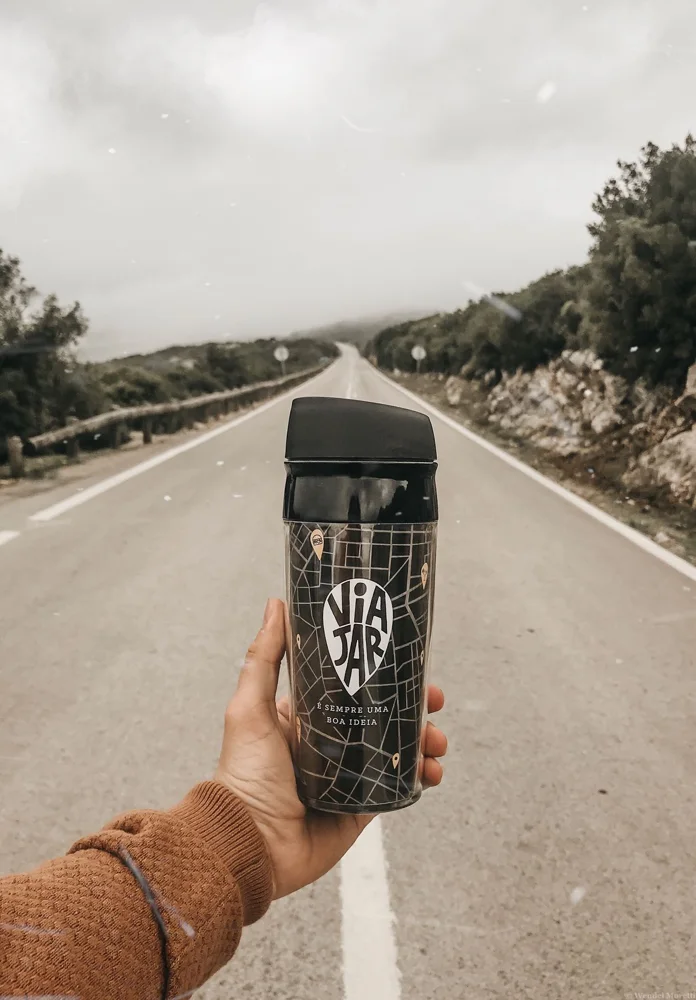 A travel mug held on an empty road surrounded by nature, suggesting adventure and travel.