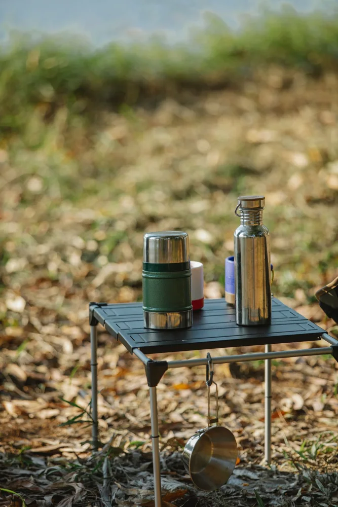 Folding table with stainless steel thermoses and mugs above saucepan on grass shore in campsite