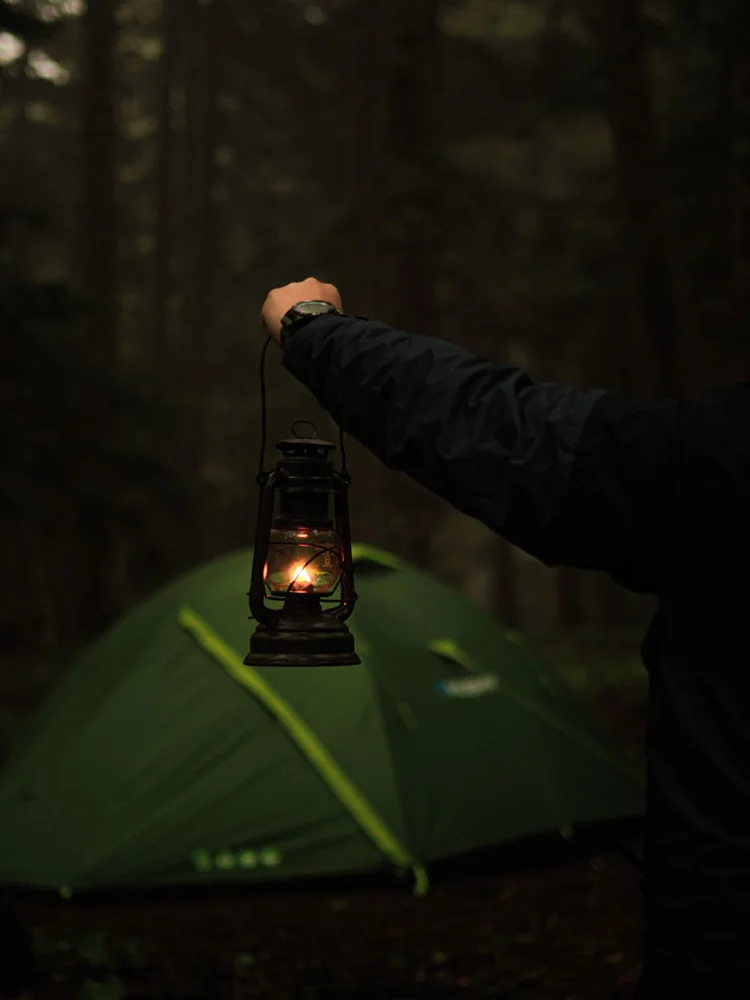 A person holds a lantern near a tent in a dark forest, capturing the essence of nighttime camping.