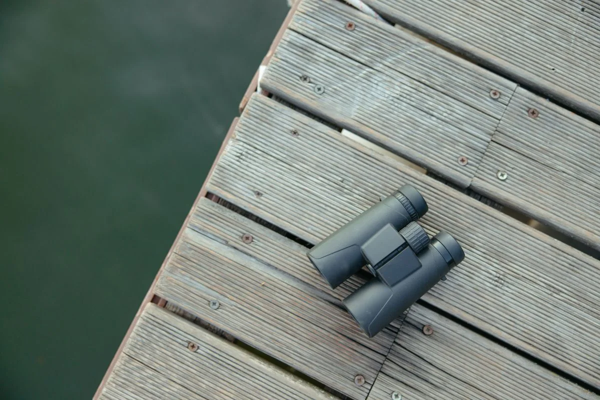 Black binoculars resting on a wooden dock with a view of water below.