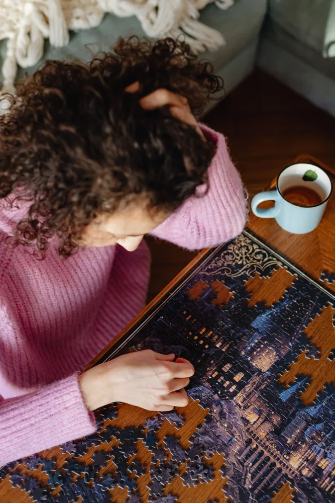 A woman in a pink sweater solves a complex puzzle at a wooden table near a cozy couch.